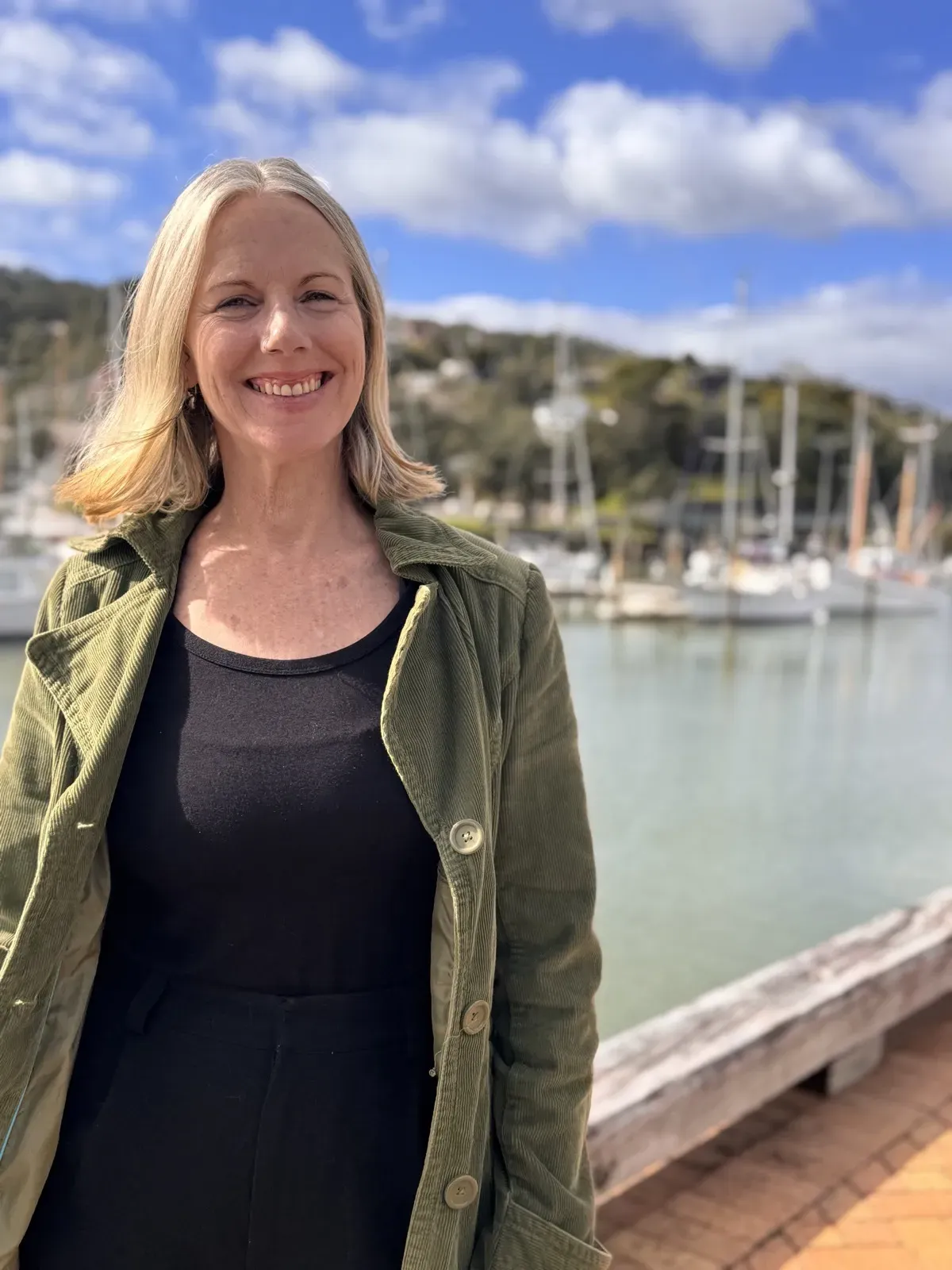 Lisa smiling at the camera in a green corduroy jacket, standing at a marina with boats and a sunny sky behind her.