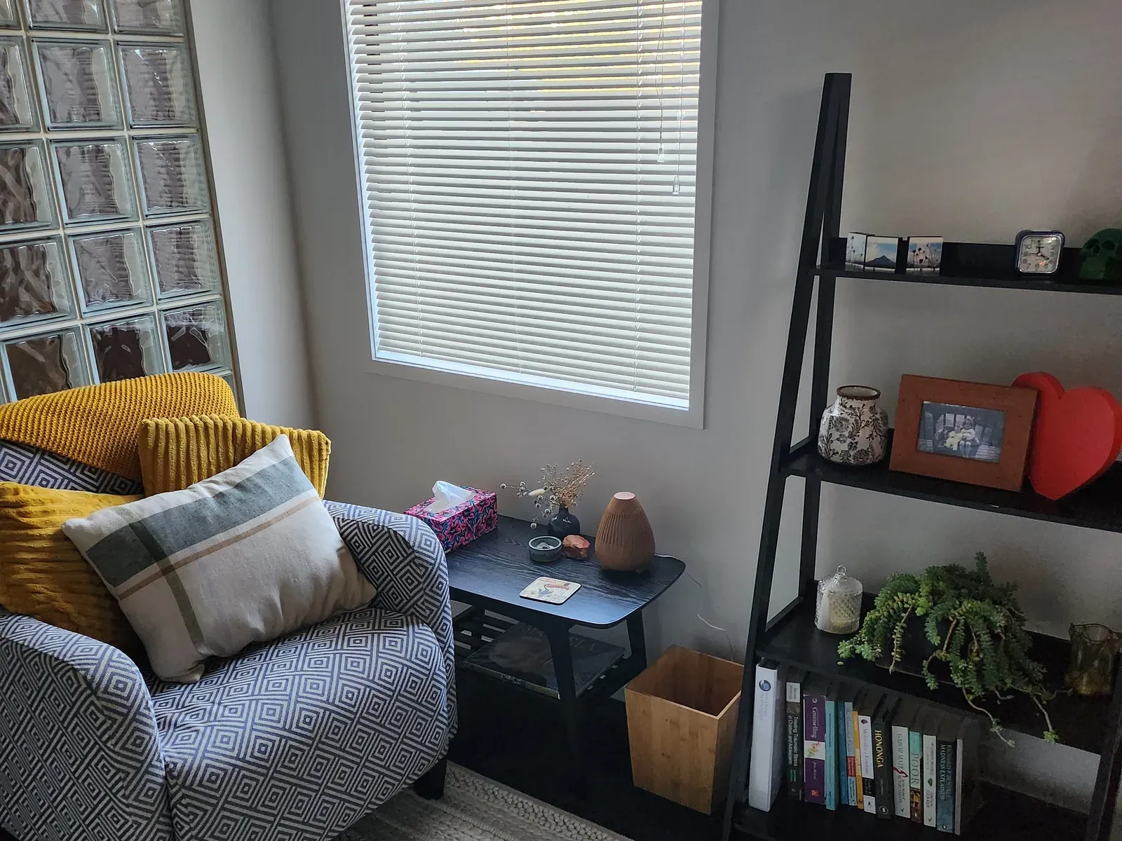 The session room: a patterned armchair with a mustard throw and cushions, a small dark side table holding tissues, a diffuser and dried flowers, and a ladder shelf of books beside a window.