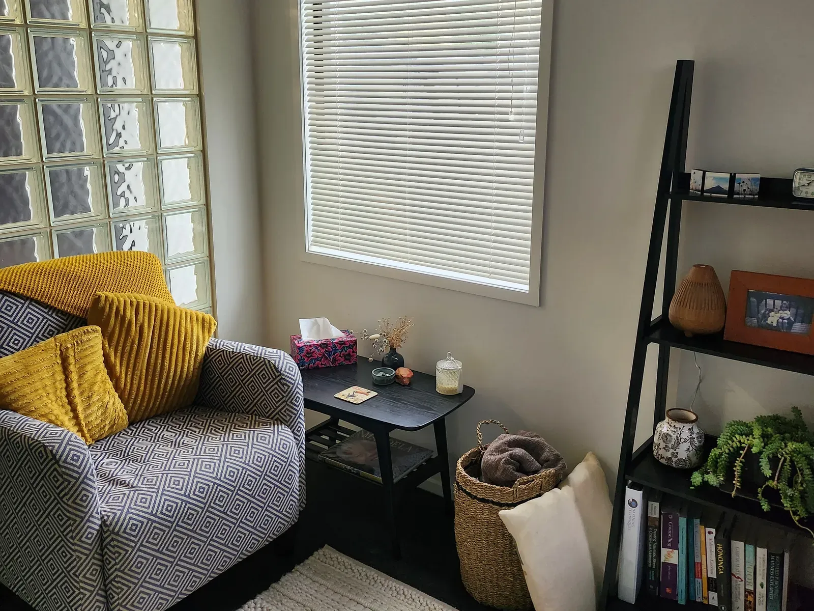 The session room seen from the doorway: a patterned armchair with mustard cushions, a side table, a basket of blankets, and daylight coming through a window and a wall of glass blocks.